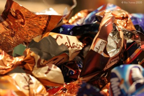 The image shows a close-up view of a pile of crumpled foil wrappers and snack packaging, primarily in metallic copper, silver, and vibrant colors with patterns and branding partially visible. The wrappers are piled together on a surface, with some edges bent and overlapping, indicating they have been discarded after use. The background is blurred but suggests an indoor setting, possibly a kitchen or a waste collection point, with warm lighting illuminating the metallic finishes of the wrappers. This scene is typical of waste materials that could be collected for rubbish removal services, such as those offered by Commercial Waste Removal Camden, especially in a residential or commercial context within the Camden area. The texture of the foil is crinkled and reflective, capturing some of the surrounding light, and the overall composition highlights a common type of household or retail waste that requires proper disposal or recycling.
