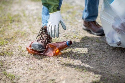 A close-up of a person's foot wearing a brown work boot with a black sole, positioned on the ground outdoors in a rural or suburban setting with patches of grass and dirt. The person is wearing blue jeans and a white glove on their left hand, which is reaching towards a discarded empty glass bottle lying on the ground. To the right, there is a partially visible large transparent plastic bag, likely containing rubbish, resting on the dirt surface. The image suggests a scene related to rubbish collection or cleanup, typical of professional waste removal services operating in Camden or nearby areas. The lighting is natural, indicating daylight, and the environment appears clean and well-maintained, reflecting a typical outdoor setting where waste removal or rubbish clearance might be undertaken by companies like Commercial Waste Removal Camden.