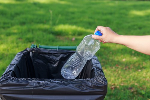 A person's hand is visible in the image, holding a clear plastic water bottle by the neck, about to place it into a black rubbish bin lined with a black plastic bag. The bin is situated outdoors on a paved surface, with neatly maintained green grass and a garden or park environment in the background. The water bottle appears mostly empty, with some residual liquid visible inside, and has a blue cap. The scene is brightly lit, suggesting natural daylight. This image reflects the importance of proper waste disposal and recycling practices, relevant to commercial waste services provided by Commercial Waste Removal Camden in Camden, London, emphasizing environmentally conscious rubbish management approach in the local area.