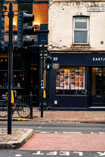 A street scene on Camden High Street featuring a historic brick building with large arched window panes and black metal accents, situated alongside a railway bridge with a large painted sign reading '