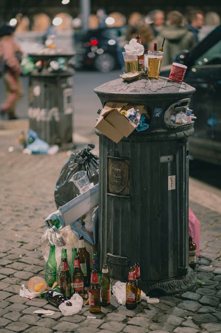 A close-up view of a pile of rubbish on the ground, including black and clear plastic garbage bags, a large yellow plastic container, and an old, dirty car tire. The debris appears to be discarded hou