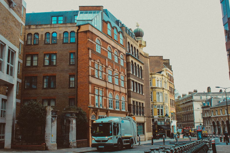 A city street scene featuring a row of multi-story buildings with varied architectural styles, including brick facades and decorative window frames. In the foreground, a white waste collection truck w